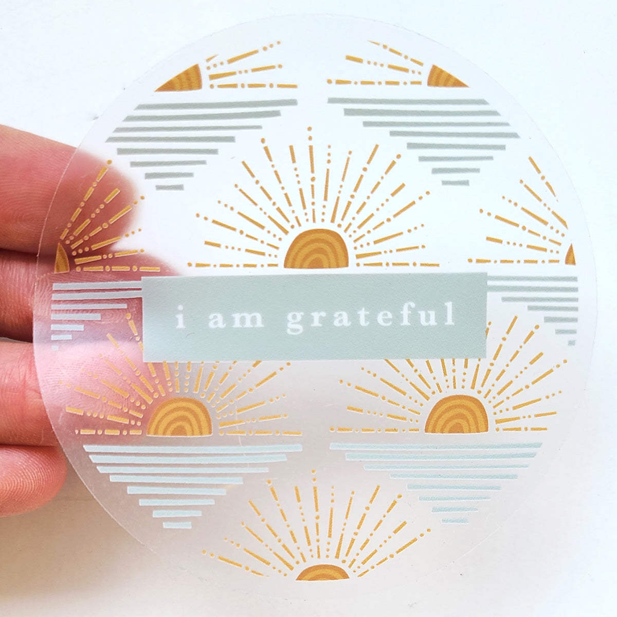 A hand holds the Sunrise 3" Clear Gratitude Sticker by Maylay Co., showcasing stylized sunrises and the phrase "I am grateful" centered amidst gold and gray sunbeam motifs on a transparent background. #PASSONTHEGRATITUDE