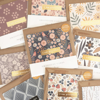 A selection of premium Baby Blooms Gratitude Greeting Cards from Maylay Co. highlights vibrant floral designs with "I am grateful" text on the packaging, elegantly arranged on a beige background.