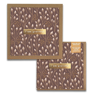 The Baby Blooms Gratitude Greeting Cards from Maylay Co. consist of two square cards with a brown background adorned with a white floral pattern. Each card features a gold strip displaying the words "I am grateful" in white text. A small label encourages you to pass on the gratitude, making these premium quality cards perfect for any occasion.