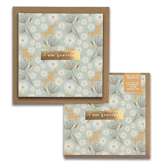 This elegant greeting card is from Maylay Co.'s Bees Floral Gratitude Greeting Cards collection and features a floral design with white and yellow flowers set against a light blue background. The words "I am grateful" are elegantly highlighted on a gold strip at the center, making it the perfect way to express your heartfelt thanks, complete with an accompanying envelope.