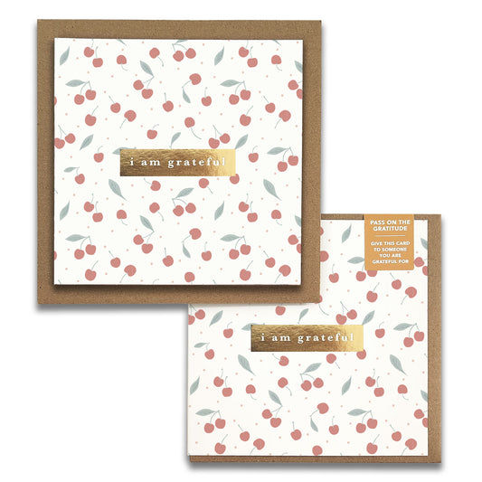 The "Cherries Gratitude Greeting Cards" by Maylay Co., featuring a cherry design and the phrase "I am grateful," are elegantly displayed on a wooden strip against a neutral background.