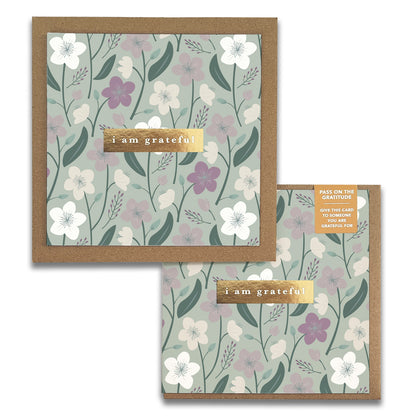 The Enchanted Garden Gratitude Greeting Cards by Maylay Co. showcase a design of white and purple flowers accompanied by green leaves on a subtle backdrop. A brown label with the message "I am grateful" is prominently featured. This thank you card set also comes with a smaller version that echoes the same design, perfect for sharing your appreciation.