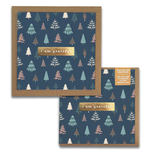 The Happy Forest Gratitude Greeting Cards by Maylay Co. feature a blue background adorned with stylized trees in green, white, and brown shades. At the center, a gold label displays "I am grateful" in white, capturing the essence of gratitude cards. A coordinating envelope is partially visible underneath.