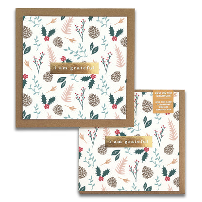 Introducing the Holiday Foli Gratitude Greeting Cards by Maylay Co. This square card boasts a festive, nature-themed design adorned with pinecones, holly leaves, and berries against a crisp white background. The card prominently features the message "I am grateful" in the center. A smaller card with an identical pattern is displayed overlapping for added appeal.