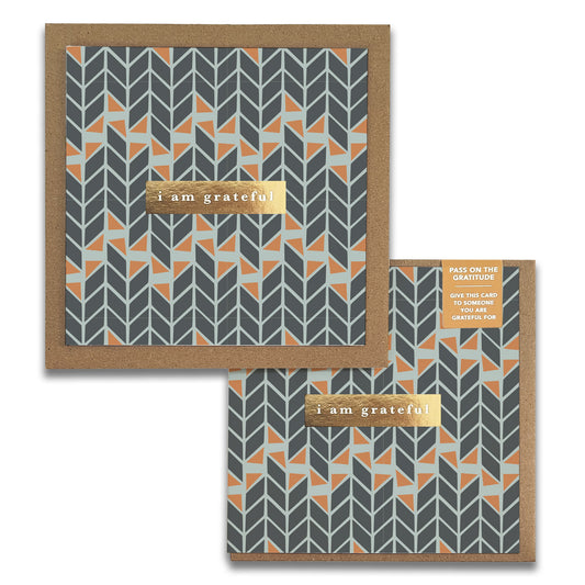 A photo of Mod Geo Gratitude Greeting Cards by Maylay Co. showcases premium quality cards with a geometric chevron pattern in dark gray, light gray, and orange. Both cards feature an elegant gold label that reads "i am grateful" and are beautifully presented in brown envelopes.