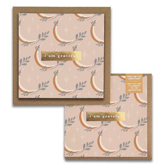 The Moonrise Gratitude Greeting Card by Maylay Co. is a square card with a beige background that displays a whimsical pattern of stylized bananas and leaves. The front is adorned with a gold label bearing the message "i am grateful" at its center. Accompanying the card is a matching envelope, ideal for sending heartfelt thanks.