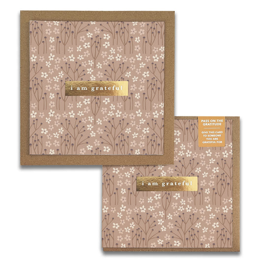 Two brown Tranquility Gratitude Greeting Cards by Maylay Co. are adorned with small white flowers and feature the phrase "I am grateful" in gold text. The note inside encourages sharing gratitude by gifting the card to someone else, with a subtle floral pattern in the background enhancing their beauty.