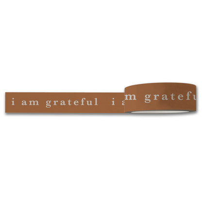 The I Am Grateful Washi Tape by Maylay Co., in a charming terracotta brown, features the phrase "i am grateful" repeatedly printed in white lowercase letters. It is partially unrolled, making it perfect for scrapbooking enthusiasts who enjoy adding a continuous personal touch of gratitude.