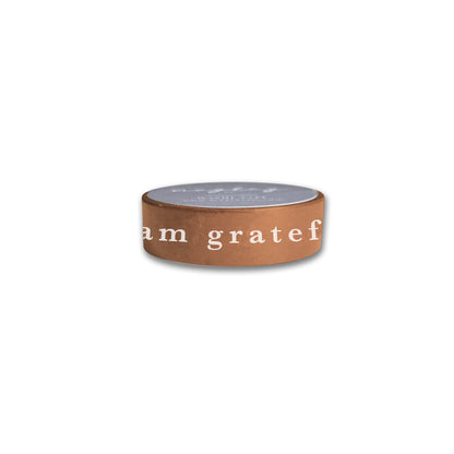 Introducing the "I Am Grateful" Washi Tape in a stylish terracotta brown from Maylay Co. This elegant tape features the inspiring phrase "I am grateful" meticulously inscribed in white letters, adding a touch of sophistication to your scrapbooking projects. Its flat gray top perfectly complements any decor and stands out beautifully against a plain white background.