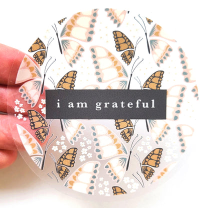 The Butterfly 3" Clear Gratitude Sticker by Maylay Co. features a design of light brown and white butterflies with small flowers on transparent vinyl. In the center, a black rectangle bears the phrase "I am grateful" in white letters. The sticker is held on its left side by a hand, making it an ideal accessory for Gratitude stickers fans to #PASSONTHEGRATITUDE.