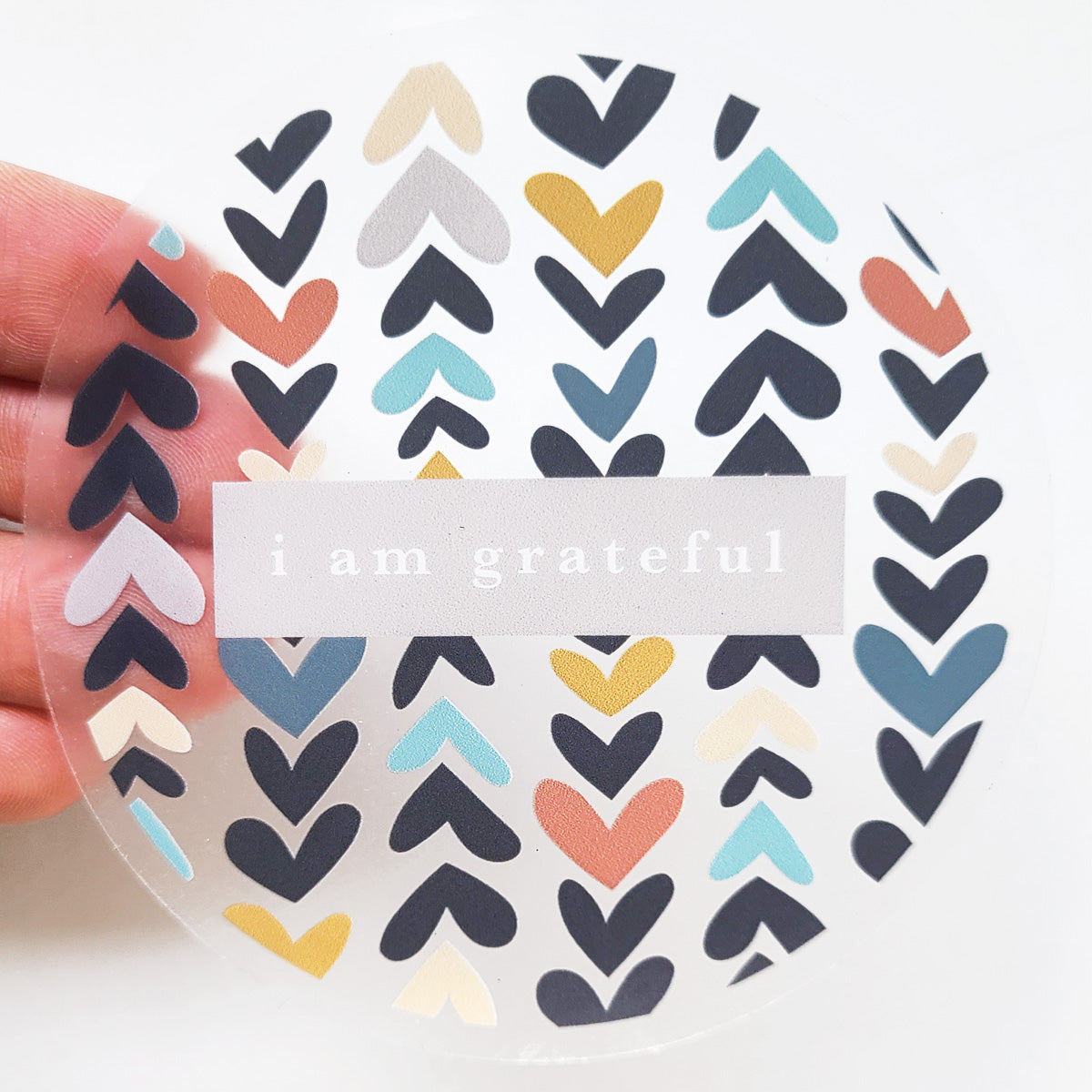 A hand holds the Heartwood 3" Clear Gratitude Sticker by Maylay Co., featuring a vibrant pattern of blue, orange, yellow, and black hearts surrounding the phrase "I am grateful" on a translucent background.