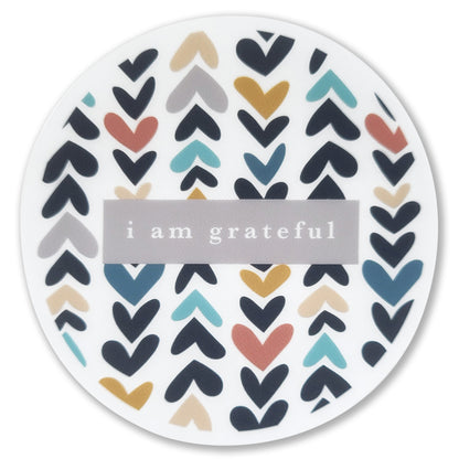 Introducing the Heartwood 3" Clear Gratitude Sticker by Maylay Co., featuring a captivating repeated pattern of vibrant hearts in black, red, blue, mustard, and gray. At its center is a gray rectangle elegantly presenting the touching message "i am grateful" in lowercase white lettering. Ideal for enthusiasts of gratitude-themed decor.