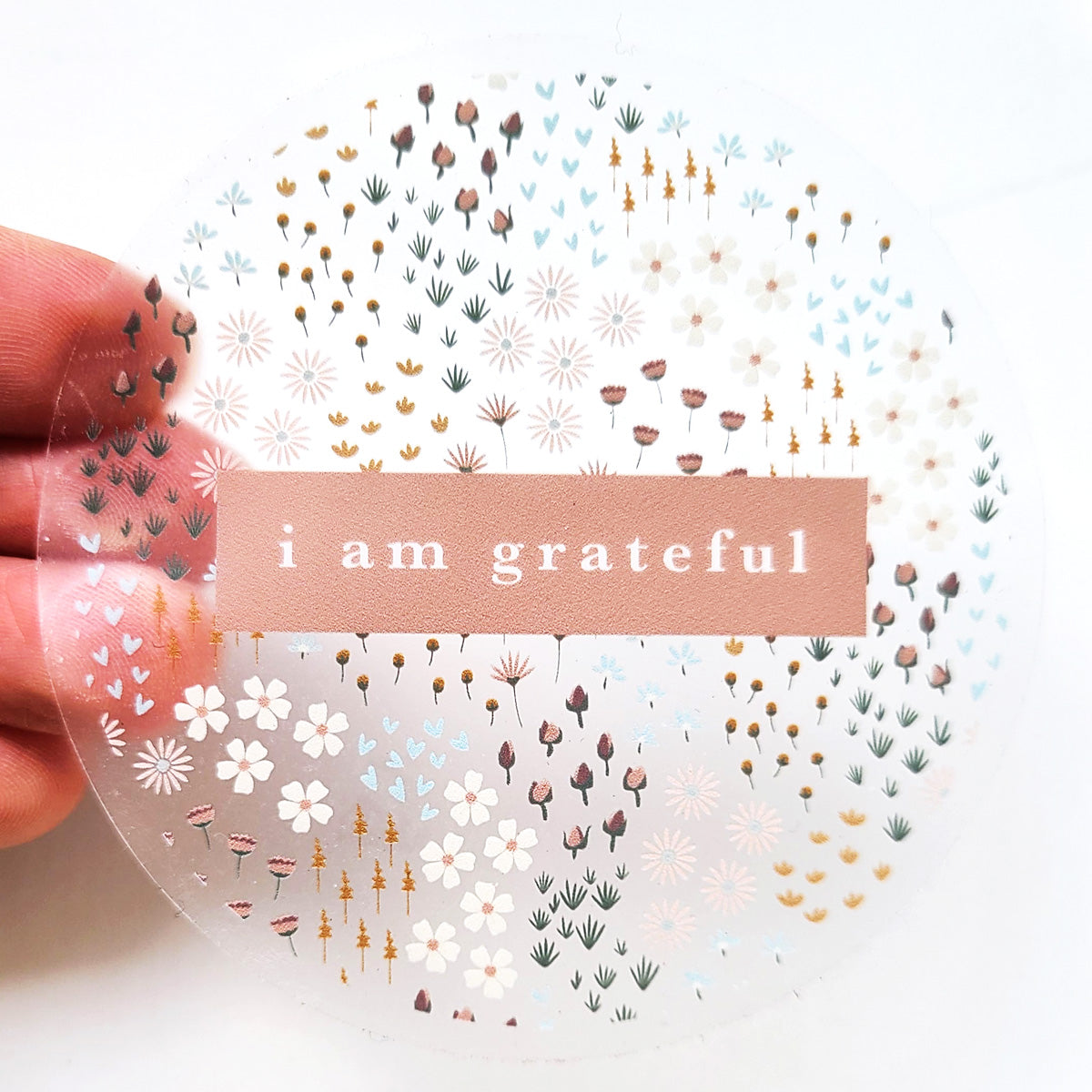 The Summerfield 3" Clear Gratitude Sticker from Maylay Co. includes a central pinkish-brown label with the phrase "i am grateful" written in white text. Surrounded by subtle floral and leaf patterns in gentle shades of white, brown, pink, and blue, it embodies timeless beauty as it's gently held by hand in this #PASSONTHEGRATITUDE piece.