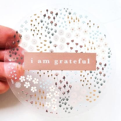 The Summerfield 3" Clear Gratitude Sticker from Maylay Co. includes a central pinkish-brown label with the phrase "i am grateful" written in white text. Surrounded by subtle floral and leaf patterns in gentle shades of white, brown, pink, and blue, it embodies timeless beauty as it's gently held by hand in this #PASSONTHEGRATITUDE piece.